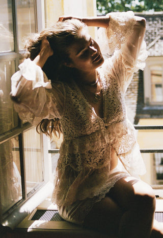 A woman wearing a sheer white lace lingerie set with ruffled sleeves and a matching sheer cover-up, sitting by a sunlit window.