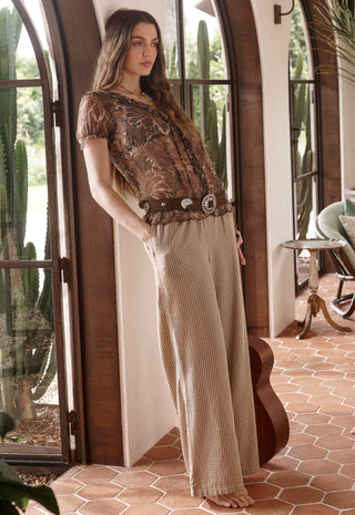A woman stands barefoot in a rustic indoor setting with arched windows and terracotta tile flooring, wearing a bohemian-style outfit consisting of a brown patterned short-sleeve top, a wide brown belt with large silver conchos, and beige wide-leg checkered pants.