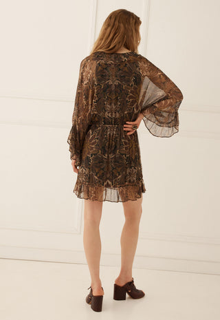 A woman viewed from behind wearing a brown sheer lace mini dress with a floral pattern and flared bell sleeves, paired with dark brown block-heel mules, set against a white paneled wall.