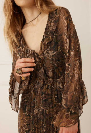 A close-up of a woman wearing a brown and black paisley-patterned sheer long-sleeve dress with a ruffled V-neckline and gathered waist.