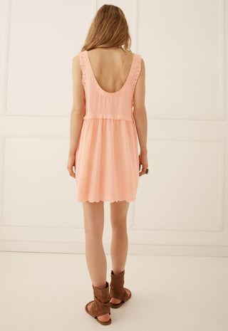 A woman viewed from behind wearing a peach-colored sleeveless dress with a ruffled back and brown leather slide sandals, standing against a white paneled wall.