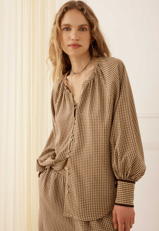 A woman wearing a brown and beige gingham long-sleeve blouse with a gathered neckline and balloon sleeves, standing against a cream-colored paneled wall.