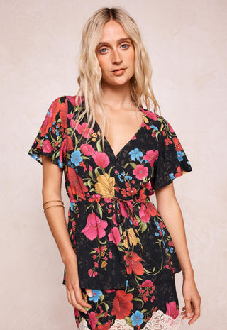 a-woman-modeling-a-black-short-sleeve-wrap-dress-with-a-vibrant-multicolor-floral-print-featuring-pink-red-blue-and-yellow-flowers-with-green-leaves-set-against-a-neutral-beige-textured-background