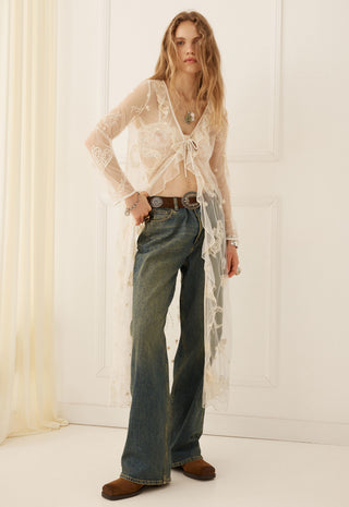 A woman stands indoors wearing a sheer white lace long-sleeve cardigan over a cropped top, blue flared jeans, and brown suede boots against a white paneled wall.